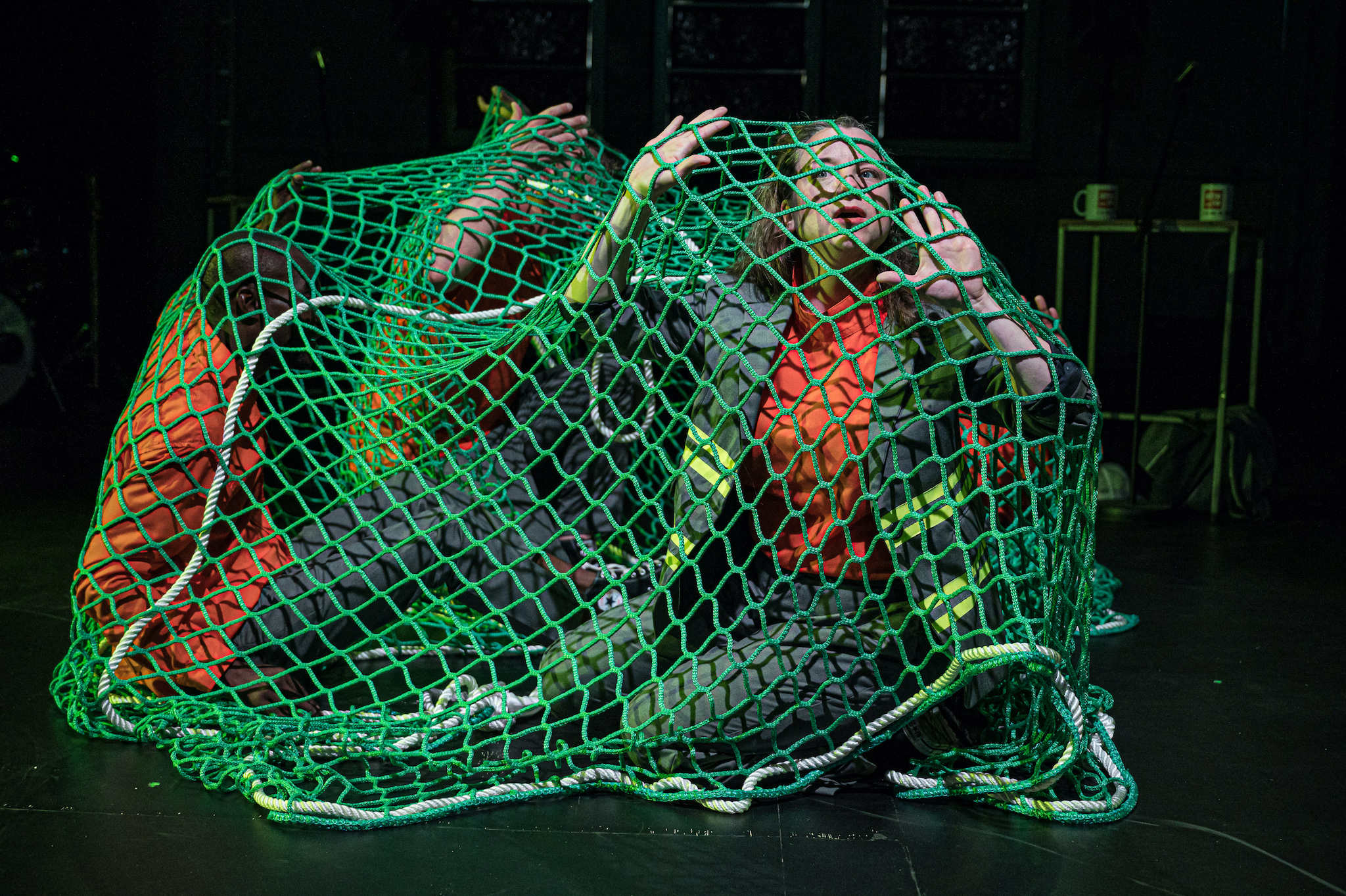 Vier Schauspieler*innen sitzen unter einem großen Netz wie gefangen.