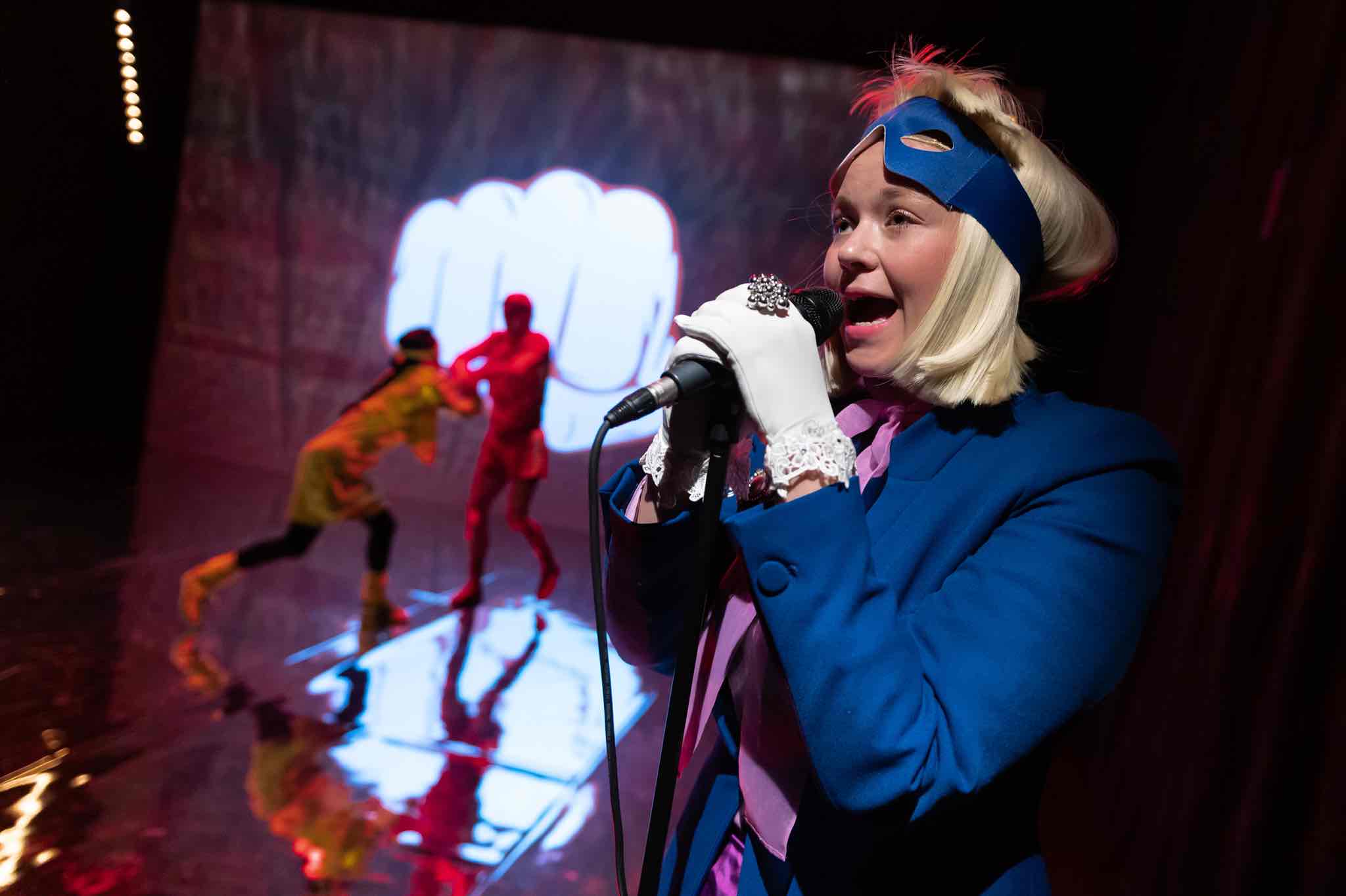 Scheitern! Schauspielerin spricht in ein Mirkofon, zwei Schauspieler*innen kämpfen im Hintergrund.